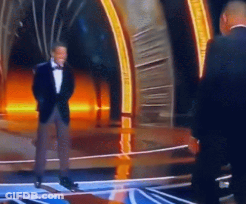 A Man In A Tuxedo Is Standing On A Stage Talking To Another Man In A Suit . GIF