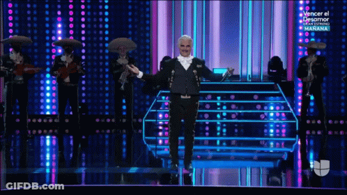 A Man In A Tuxedo Is Standing On A Stage With Mariachi Players Behind Him GIF