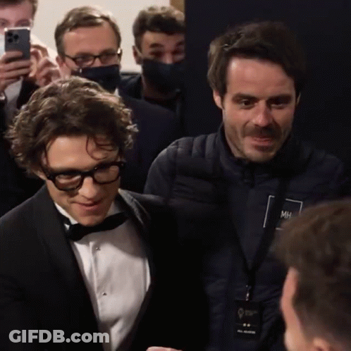 A Man In A Tuxedo Is Talking To Another Man In A Jacket With Mh On It GIF