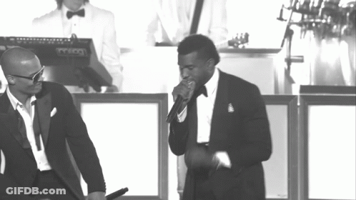 A Man In A Tuxedo Sings Into A Microphone While Another Man Stands Behind Him Playing A Keyboard GIF