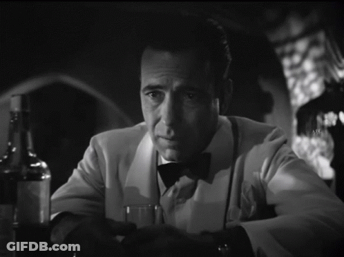 A Man In A Tuxedo Sits At A Table With A Glass And A Bottle Of Alcohol GIF
