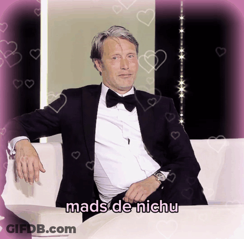 A Man In A Tuxedo Sits On A White Couch With The Words Mads De Nichu Written Below Him GIF