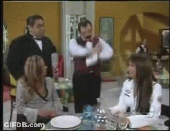 A Man In A Tuxedo Stands Behind A Woman Sitting At A Table With Plates And Wine Glasses GIF