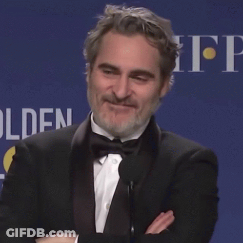 A Man In A Tuxedo Stands In Front Of A Microphone With His Arms Crossed In Front Of A Sign That Says Golden GIF