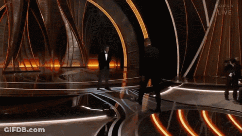 A Man In A Tuxedo Stands On A Stage With The Word Live On The Bottom Left GIF