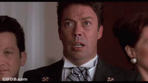 A Man In A Tuxedo With A Bow Tie Looks At The Camera With A Woman Behind Him GIF