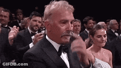 A Man In A Tuxedo With A Mustache Is Applauding In A Crowd . GIF