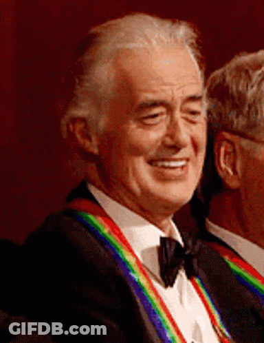 A Man In A Tuxedo With A Rainbow Ribbon Around His Neck Is Smiling GIF