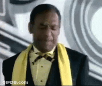 A Man In A Tuxedo With A Yellow Scarf Around His Neck Is Making A Face . GIF
