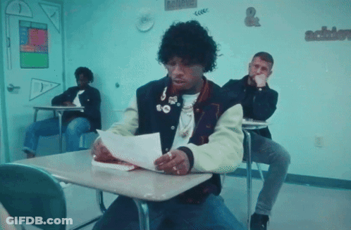 A Man In A Varsity Jacket Is Sitting At A Desk Reading A Piece Of Paper GIF