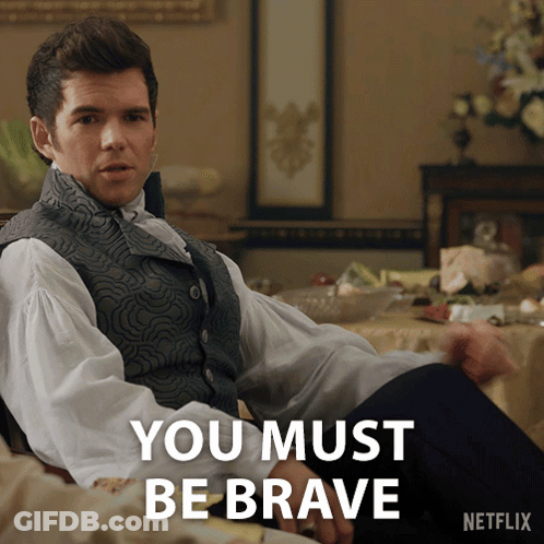 A Man In A Vest Sits In Front Of A Table With The Words " You Must Be Brave " On It GIF