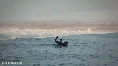A Man In A Wetsuit Is Floating In The Ocean With His Hand Up GIF