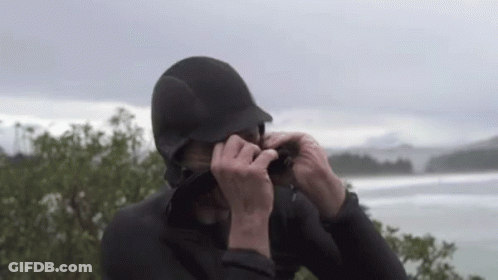 A Man In A Wetsuit Is Taking A Picture Of A Body Of Water . GIF
