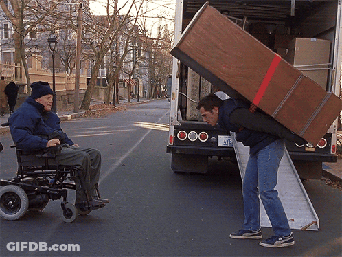 A Man In A Wheelchair Is Being Helped By A Man Carrying A Large Wooden Box GIF