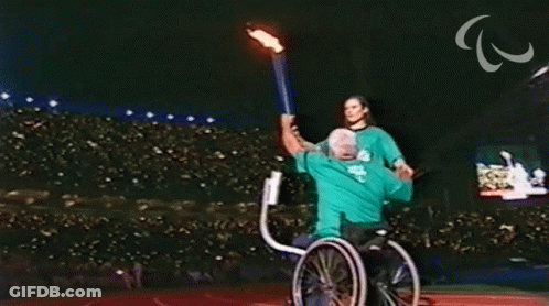 A Man In A Wheelchair Is Being Helped By A Woman Holding A Fire Torch GIF