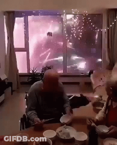 A Man In A Wheelchair Sits At A Table With Bowls And Plates In Front Of A Window Watching Fireworks GIF