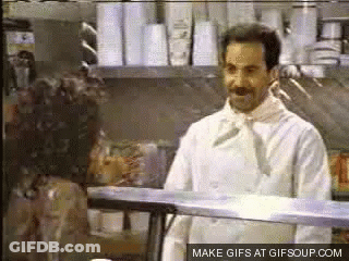 A Man In A White Chef 'S Coat Is Standing In Front Of A Display Case . GIF