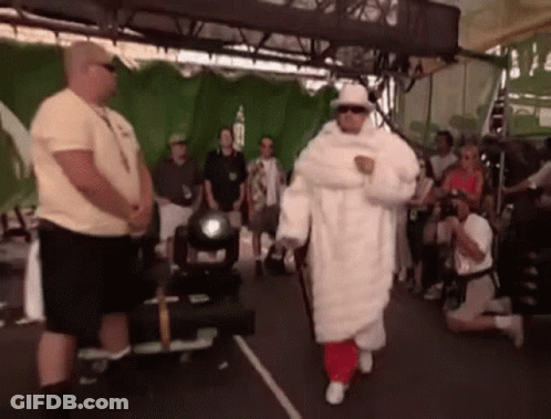 A Man In A White Fur Coat And Hat Is Walking In Front Of A Crowd . GIF