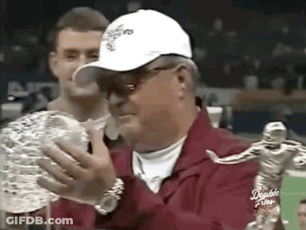 A Man In A White Hat Is Holding A Trophy With A Statue Of A Football Player In The Background . GIF