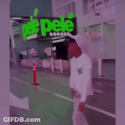 A Man In A White Hat Is Standing In Front Of A Building With A Green Sign That Says Soccer . GIF