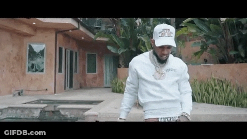 A Man In A White Hat Is Standing In Front Of A Large House . GIF