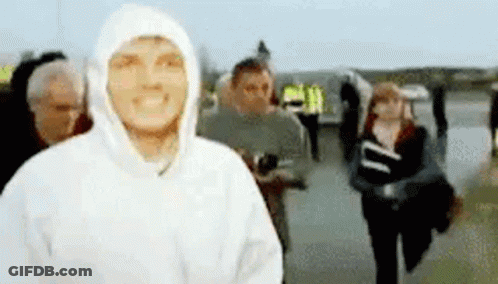 A Man In A White Hoodie Is Smiling In Front Of A Crowd Of People . GIF