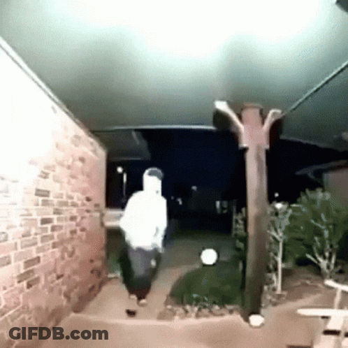A Man In A White Hoodie Is Walking Down A Sidewalk Next To A Brick Wall . GIF