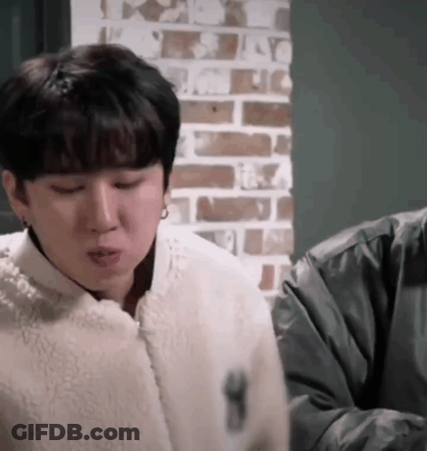 A Man In A White Jacket Is Sitting In Front Of A Brick Wall And Making A Funny Face . GIF