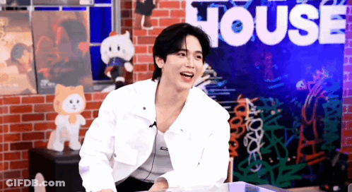 A Man In A White Jacket Is Sitting In Front Of A Brick Wall With The Word House On It . GIF