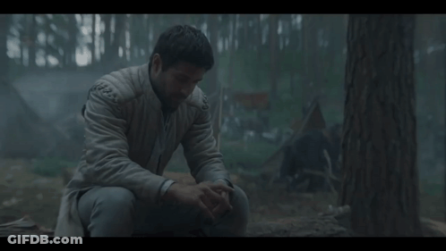 A Man In A White Jacket Sits In The Woods With His Hands On His Knees GIF