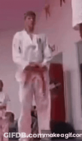 A Man In A White Karate Uniform Is Standing In A Room With His Belt Around His Waist . GIF