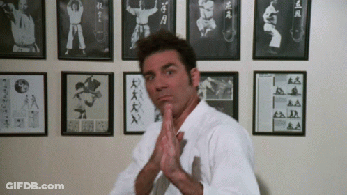 A Man In A White Karate Uniform Stands In Front Of A Wall Of Framed Pictures Of Karate Fighters GIF