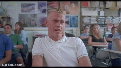 A Man In A White Polo Shirt Sits At A Desk In A Classroom GIF