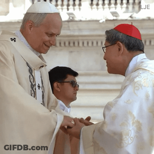 A Man In A White Robe Is Shaking Hands With A Man Wearing A Red Hat GIF