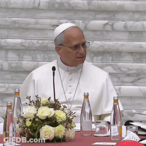 A Man In A White Robe Is Sitting At A Table With Bottles Of Water And Flowers GIF
