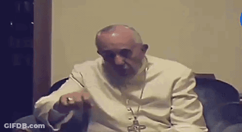 A Man In A White Robe Is Sitting In A Chair With A Cross On His Neck . GIF