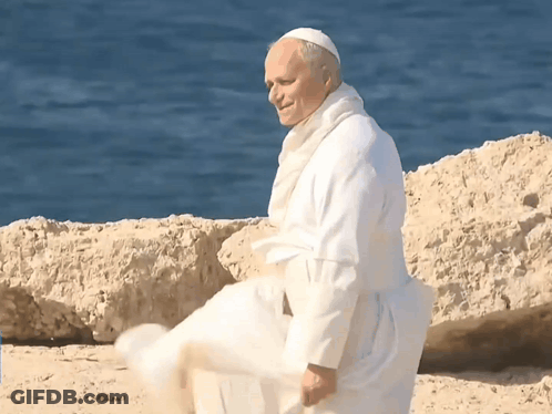 A Man In A White Robe Is Standing On A Beach Near The Water GIF