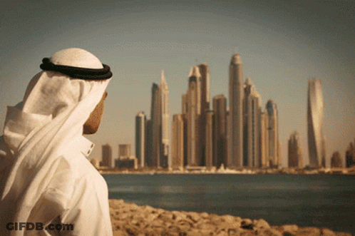 A Man In A White Robe Looks Out Over A Body Of Water At A City Skyline GIF