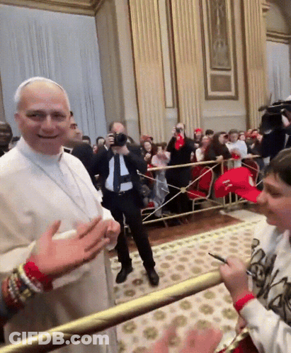 A Man In A White Robe Stands In Front Of A Crowd While A Boy In A Red Hat Stands Behind Him GIF