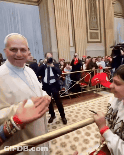 A Man In A White Robe Stands In Front Of A Crowd While A Boy Wearing A Red Hat With The Letter S On It GIF