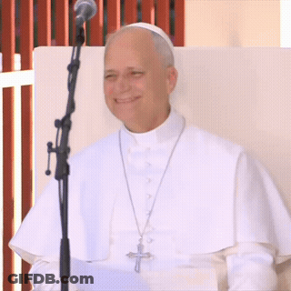 A Man In A White Robe Stands In Front Of A Microphone With A Cross On His Necklace GIF