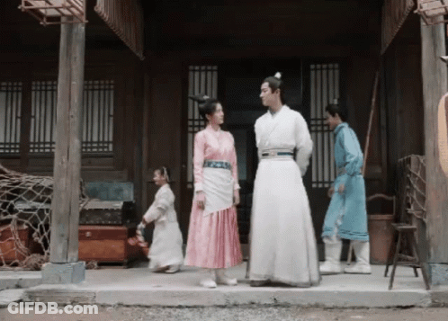 A Man In A White Robe Stands Next To A Woman In A Pink Dress On A Porch GIF