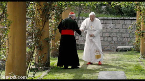 A Man In A White Robe With A Cross On It Stands Next To Another Man In A Black Robe GIF