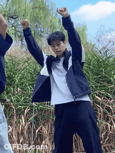 A Man In A White Shirt And A Black Jacket Is Standing In Front Of Tall Grass GIF