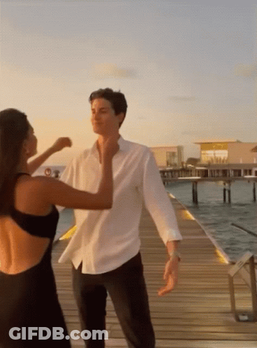 A Man In A White Shirt And A Woman In A Black Dress Are Dancing On A Dock GIF