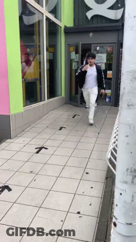 Man In White Shirt And Black Jacket Walking Down Sidewalk GIF
