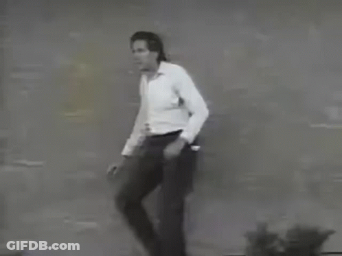 A Man In A White Shirt And Black Pants Is Standing In Front Of A Brick Wall . GIF