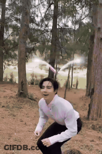 A Man In A White Shirt And Black Pants Squats In The Woods GIF