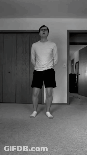 A Man In A White Shirt And Black Shorts Is Standing In A Living Room GIF