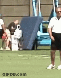 A Man In A White Shirt And Black Shorts Is Standing On A Green Field . GIF